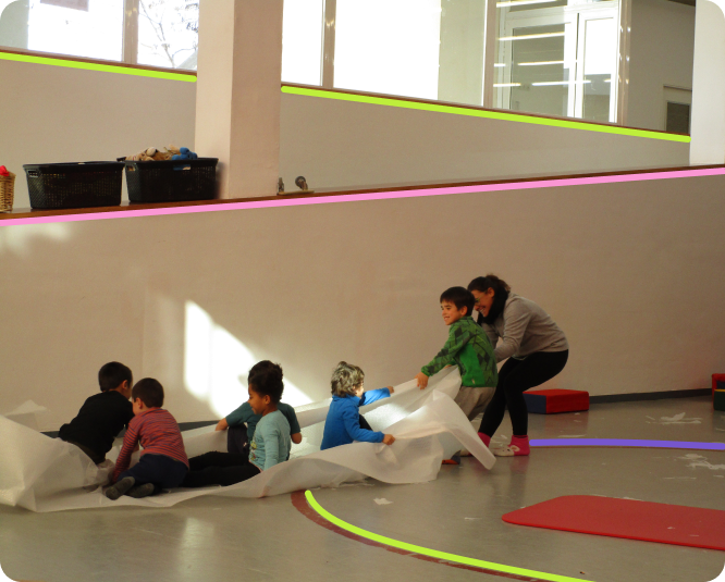 A child, assisted by an adult, is drawing a large piece of fabric with five other children seated on it.