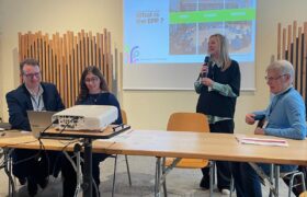 Three persons sitting while a fourth one is standinf presenting a powerpoint